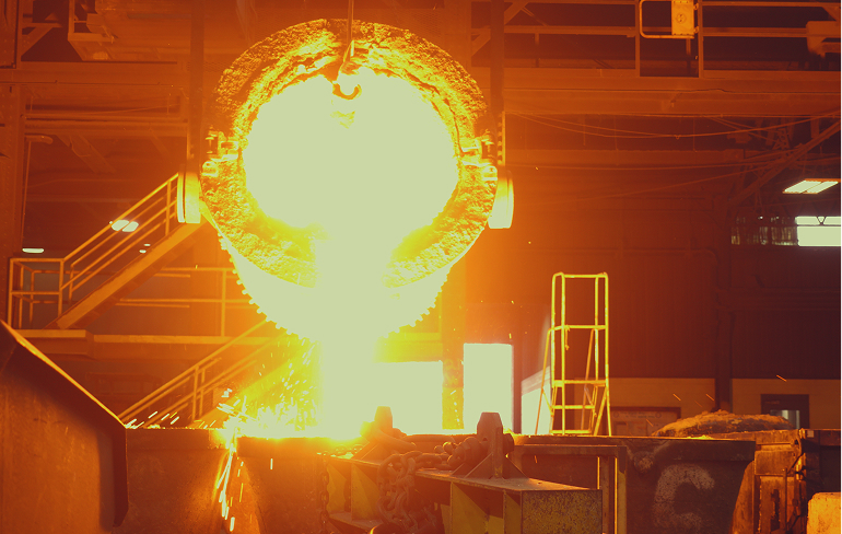 Dramatic industrial scene showing molten metal being poured from a large crucible, creating a brilliant orange-yellow stream of liquid steel flowing into a casting mold. The intense heat illuminates the surrounding foundry environment during this critical manufacturing process