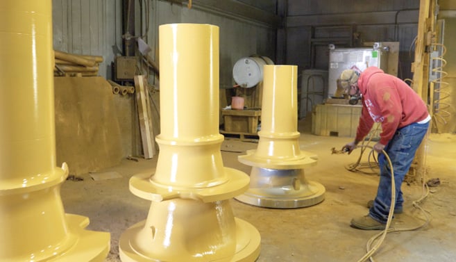 Industrial worker applying bright yellow protective coating to a massive finished steel casting, highlighting the final stage of the manufacturing process before the large metal component is ready for installation or shipment.