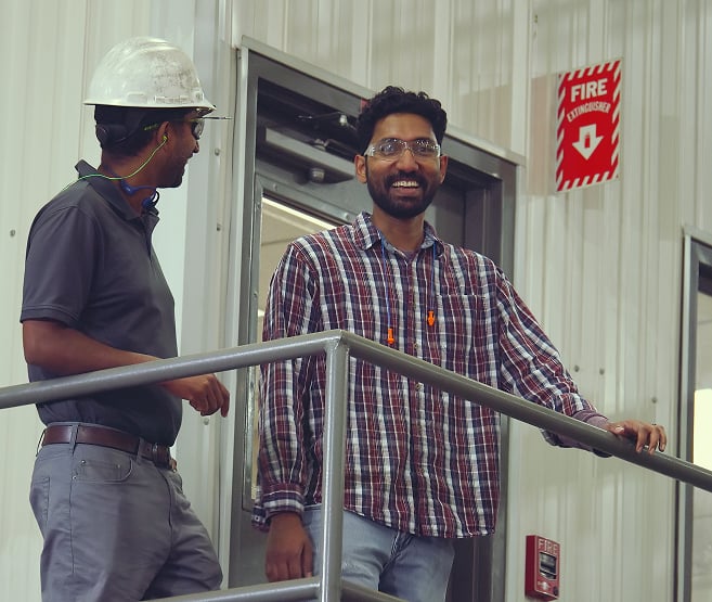 Two employees, smiling, stand on top of the stairs next o an office entrance. A fire alarm is visible in the background.