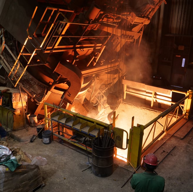 Industrial casting equipment elevated to reveal its intense orange-glowing interior chamber where molten metal is heated to extreme temperatures before being poured into molds for large-scale steel castings."